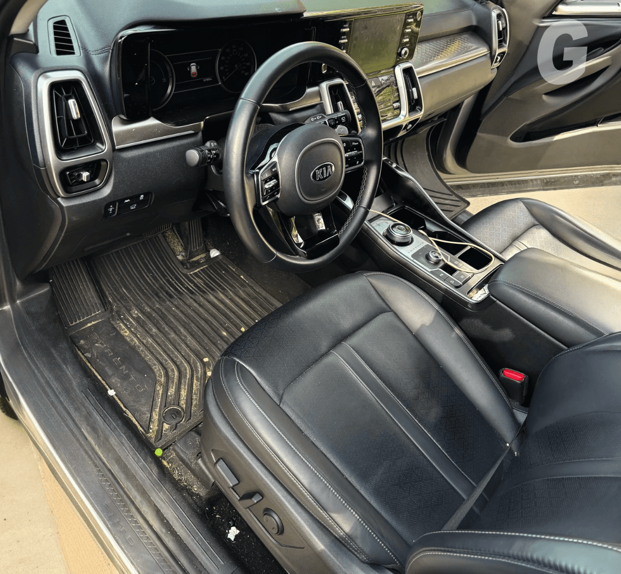 Front interior of a Kia with black leather seats and a rubber floor mat covered in dirt and crumbs, driver door open