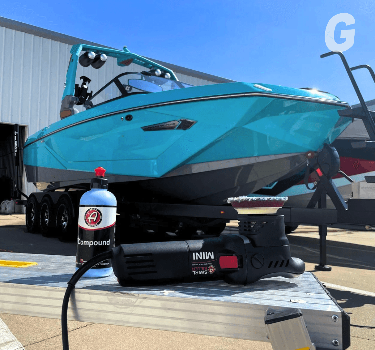 Turquoise wake boat on a trailer with a bottle of compound and a mini polisher resting on a metal ramp in the sun