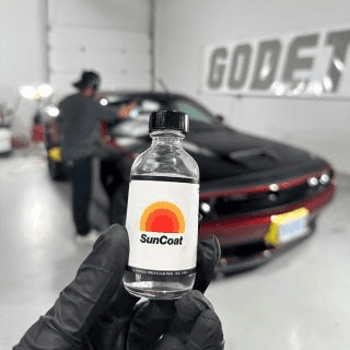 Gloved hand holding a small SunCoat bottle in a shop while a person works on a dark red sports car behind it