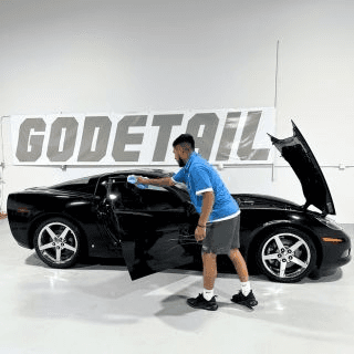 Detailer in a blue shirt wipes the open door of a black sports car in a workshop with a GoDetail banner behind it