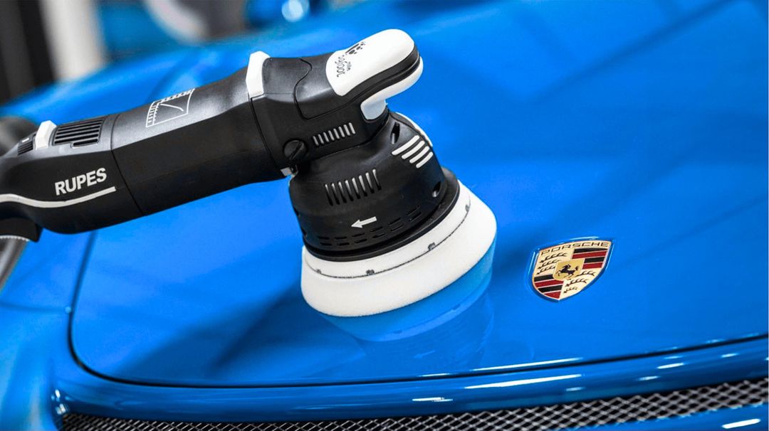 Rupes machine polisher with a white foam pad buffing the glossy blue hood of a Porsche near its crest badge