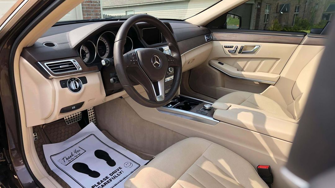 Clean cream leather interior of a Mercedes with dark wood trim, steering wheel and a paper floor mat on the driver side