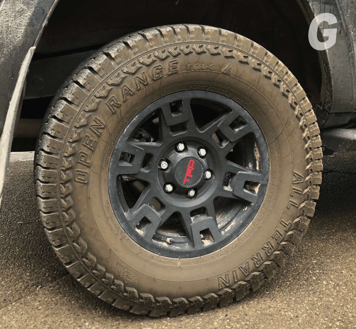 Mud-caked all terrain tire on a black wheel with a red TRD center cap, on a truck parked on wet pavement