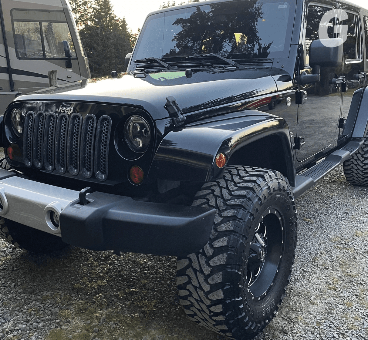 Glossy black Jeep Wrangler with large off-road tires parked on gravel next to a motorhome, trees in the background
