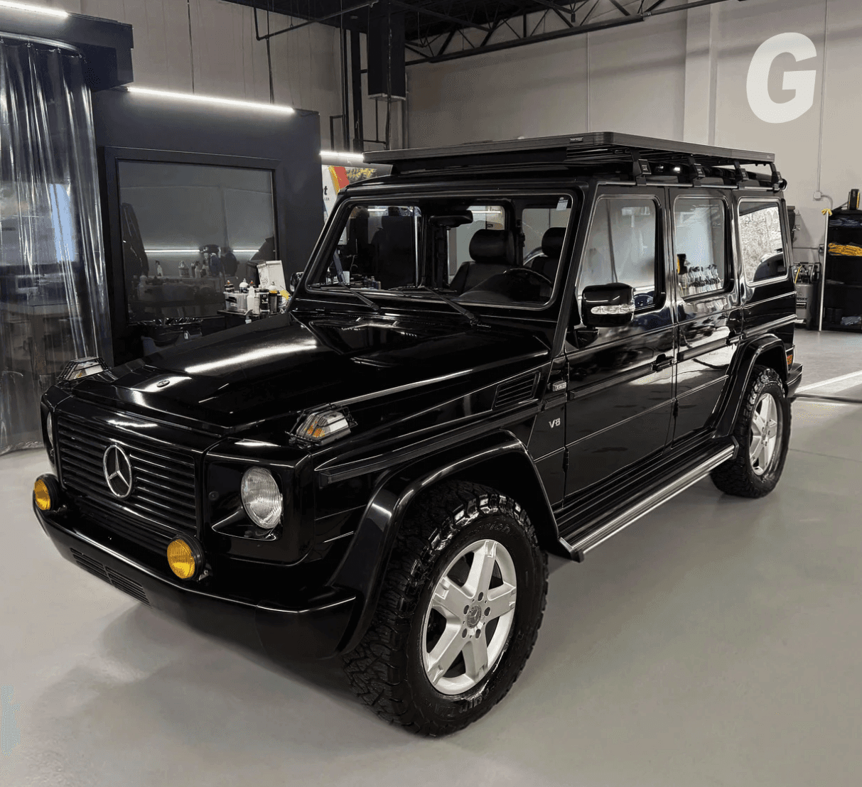 Glossy black Mercedes G-Class SUV with a roof rack and silver wheels parked inside a clean, bright workshop
