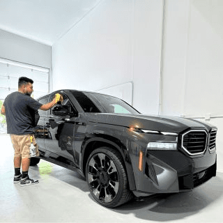 Man in a gray t-shirt and khaki shorts polishes the side of a dark gray SUV inside a bright white garage