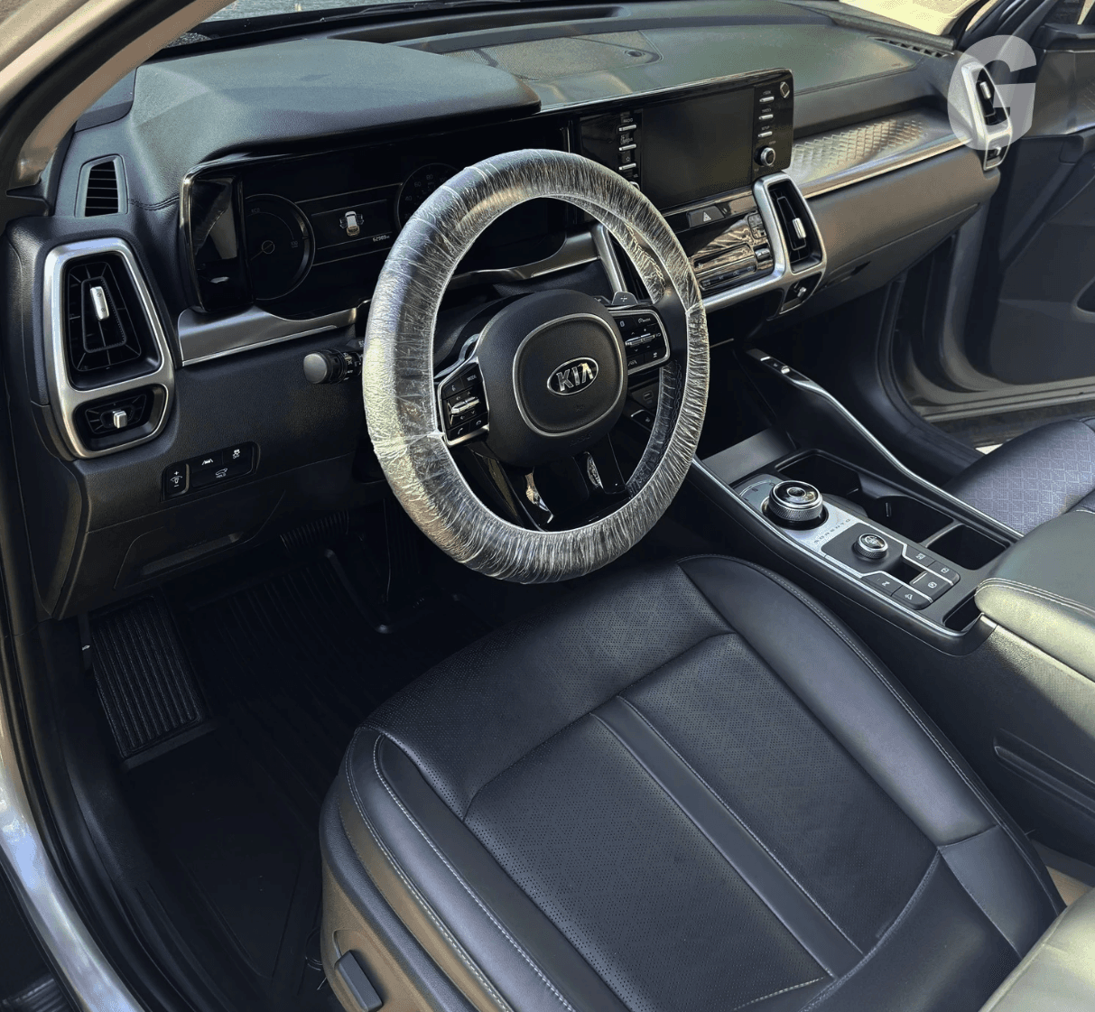 Clean black leather interior of a Kia with a plastic cover wrapped around the steering wheel and rubber floor mats