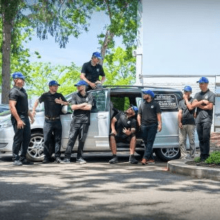 Group of eight men in black shirts and blue caps posing around a silver van with its side door open, one standing on the roof