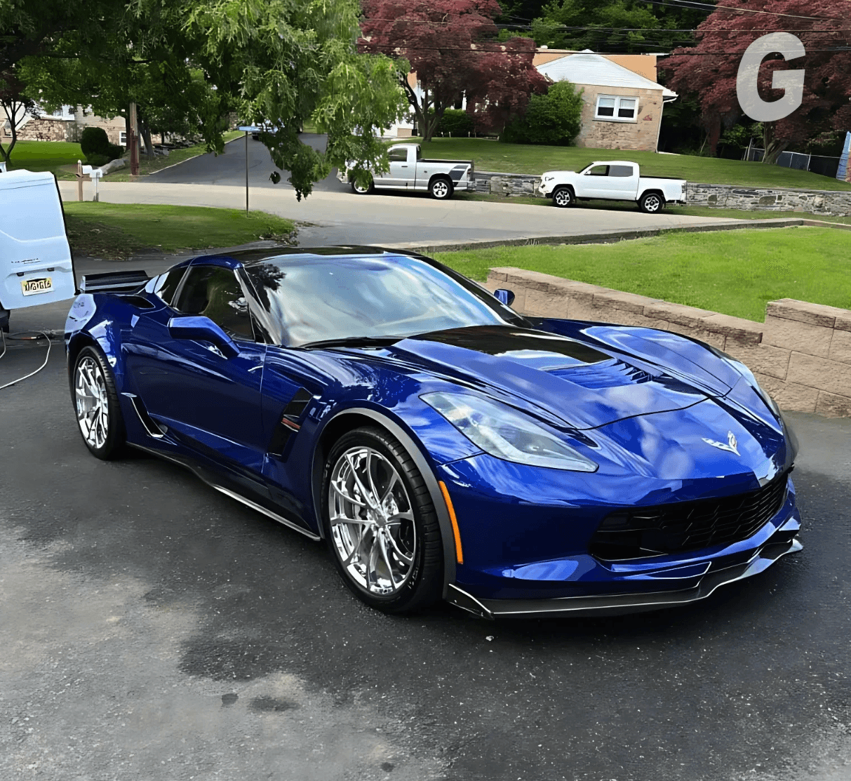 Glossy dark blue sports car with chrome wheels parked on a wet driveway next to a white van, with houses and trees behind
