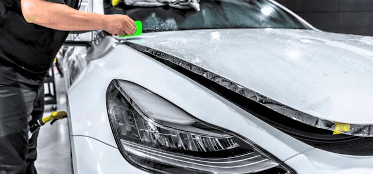 Worker in a black shirt smooths wet clear film on the hood of a white car with a green squeegee in a shop