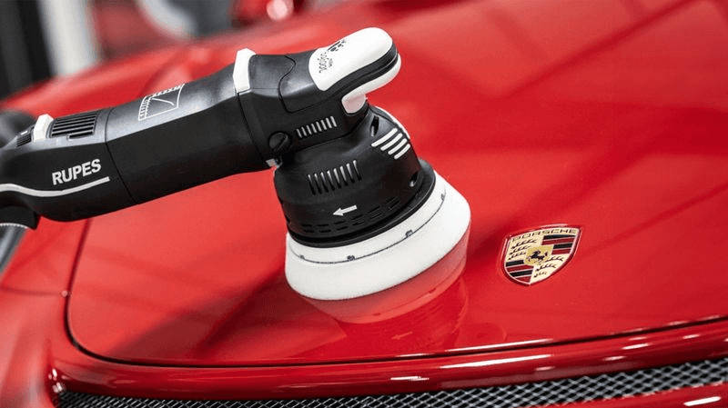 Rupes machine polisher with a white foam pad buffing the glossy red hood of a Porsche