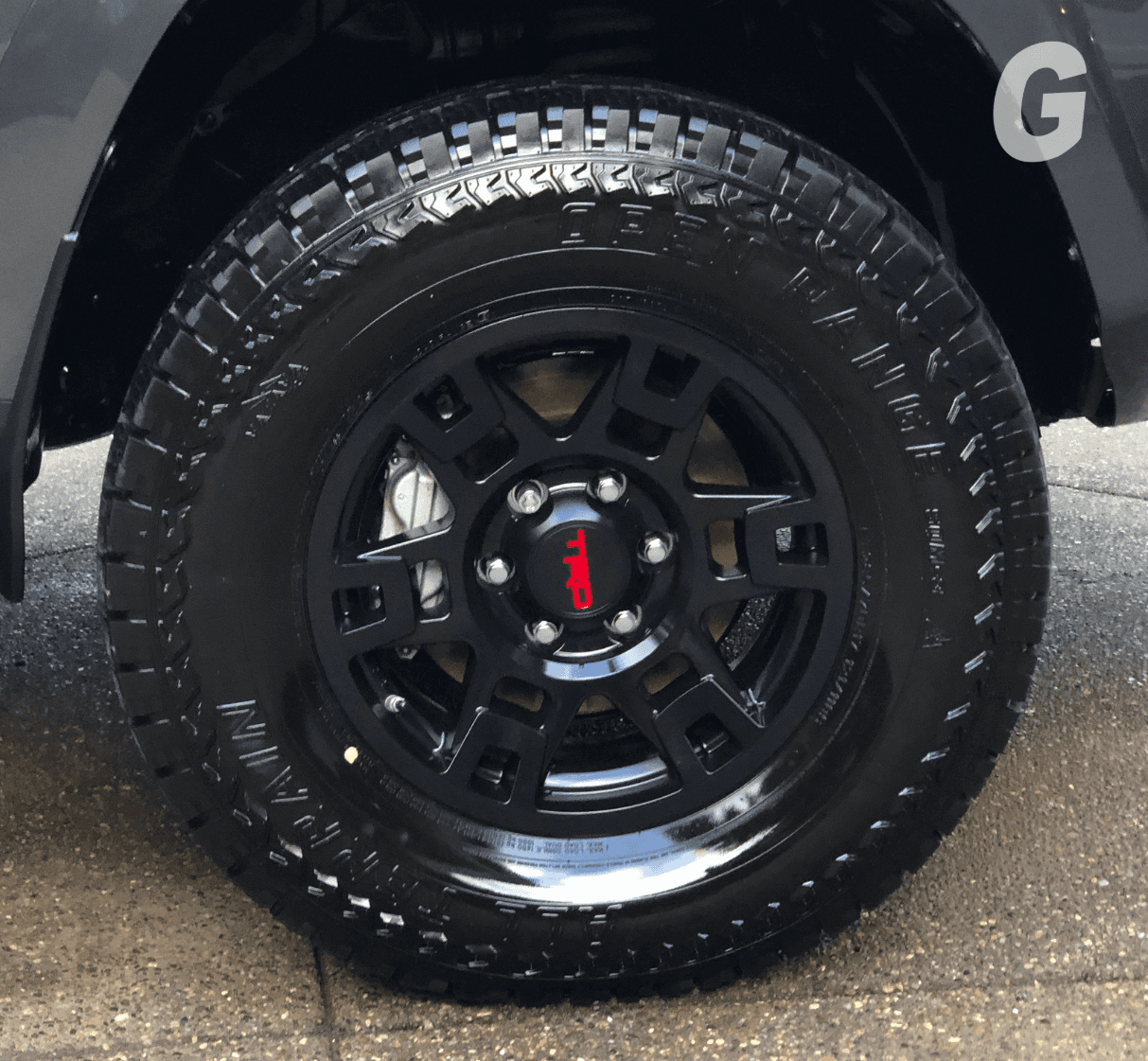 Close view of a glossy black truck tire and black wheel with a red TRD center cap, parked on wet pavement