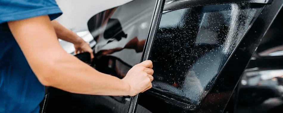 Person in a blue shirt holding the edge of a wet, dark tinted car window while working on it