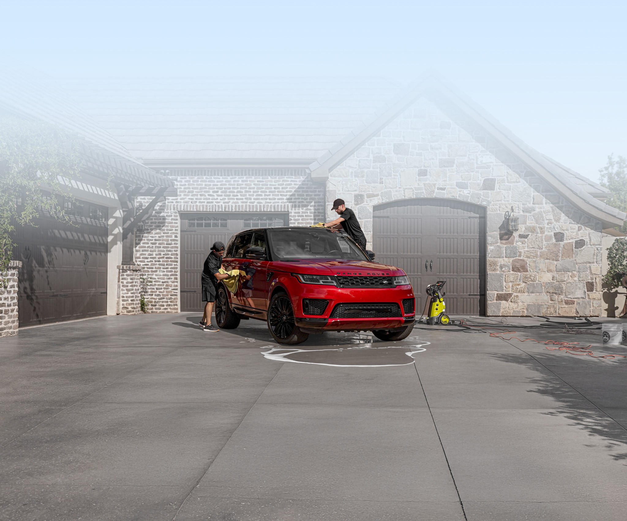 Two men in black wipe down a red SUV with yellow towels on a driveway in front of a stone and brick house with garages