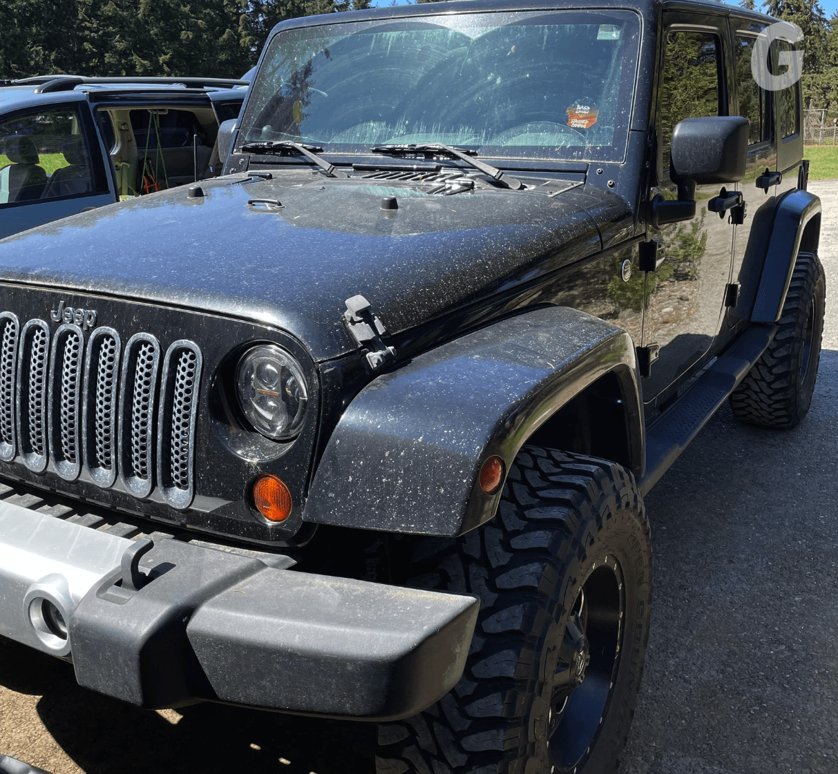 Front of a black Jeep Wrangler covered in dust and spots, with large off-road tires, parked on a gravel lot