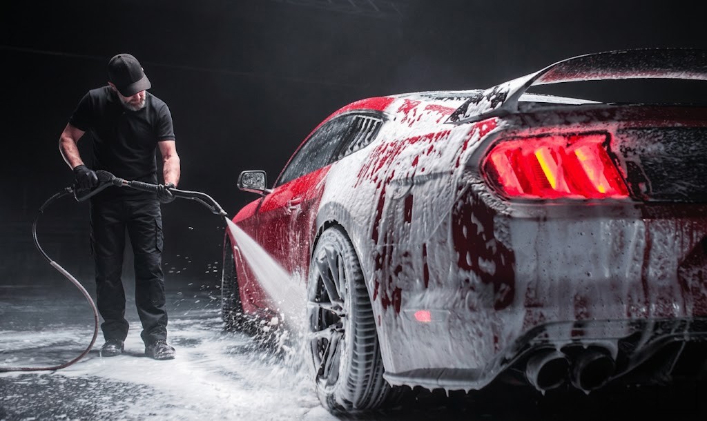 Man in a black cap and gloves rinses a red sports car covered in thick white foam with a pressure washer