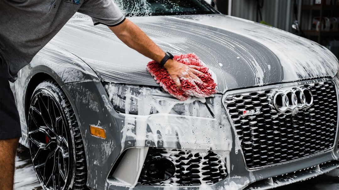 Detailer scrubbing a soapy gray Audi hood with a red microfiber wash mitt, black wheel and honeycomb grille in view